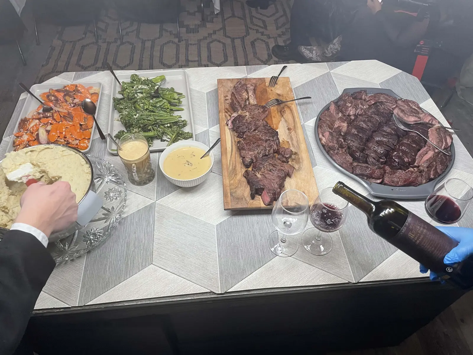 A catered table spread with sliced steak, roasted carrots, broccolini and wine being poured